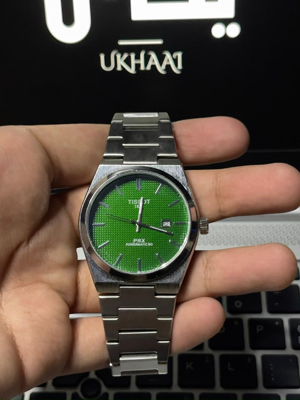 Hand holding a silver watch with a green face against a blurred background