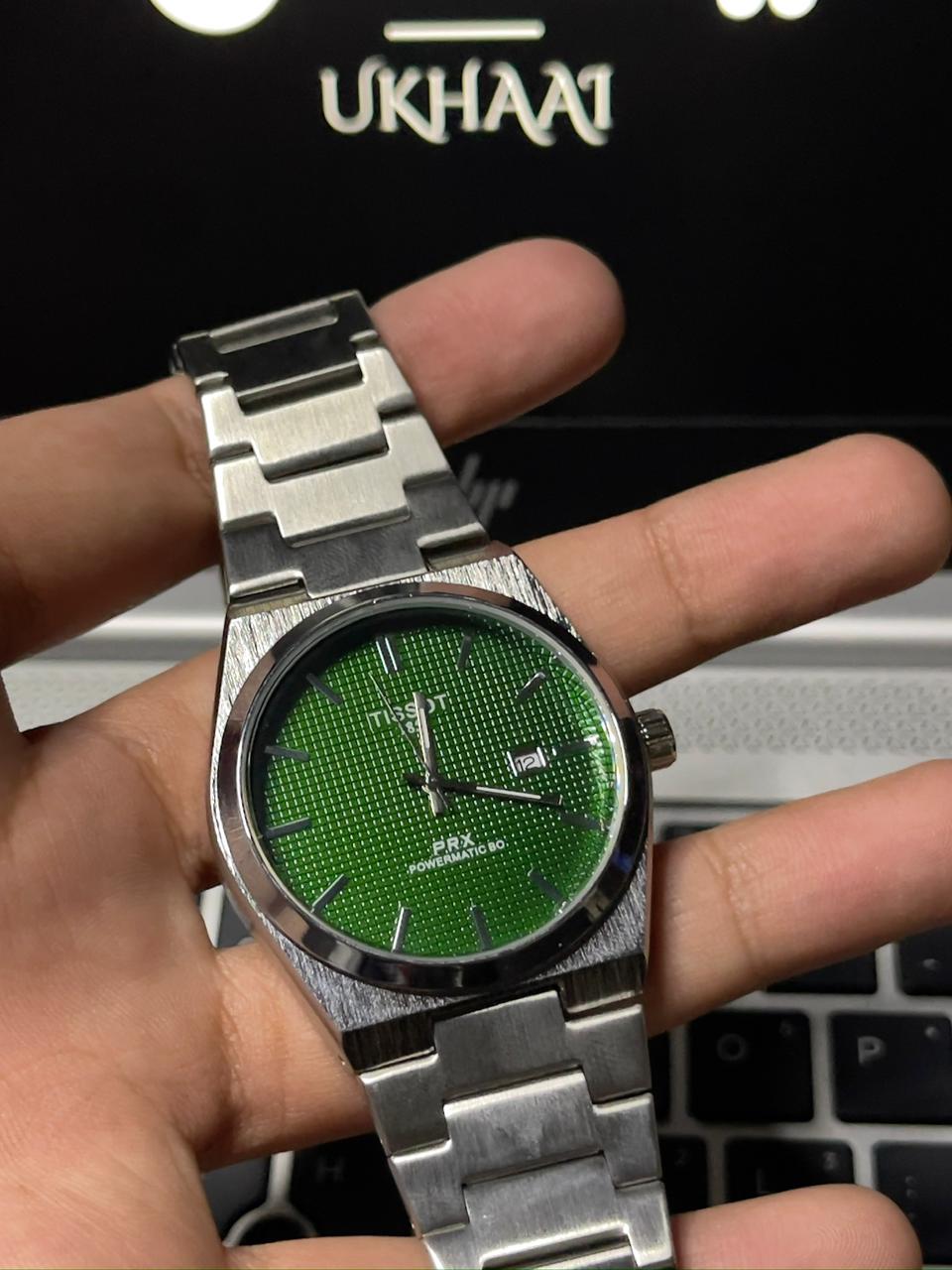 Hand holding a silver watch with a green dial against a blurred background
