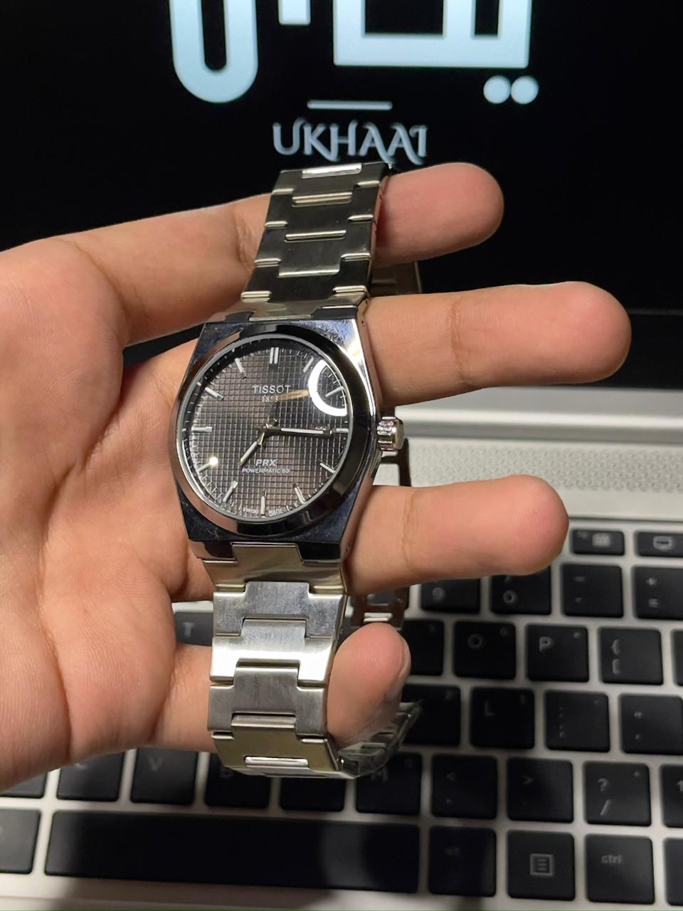 Hand holding a silver watch in front of a laptop with 'UKHAAI' branding.(front side)