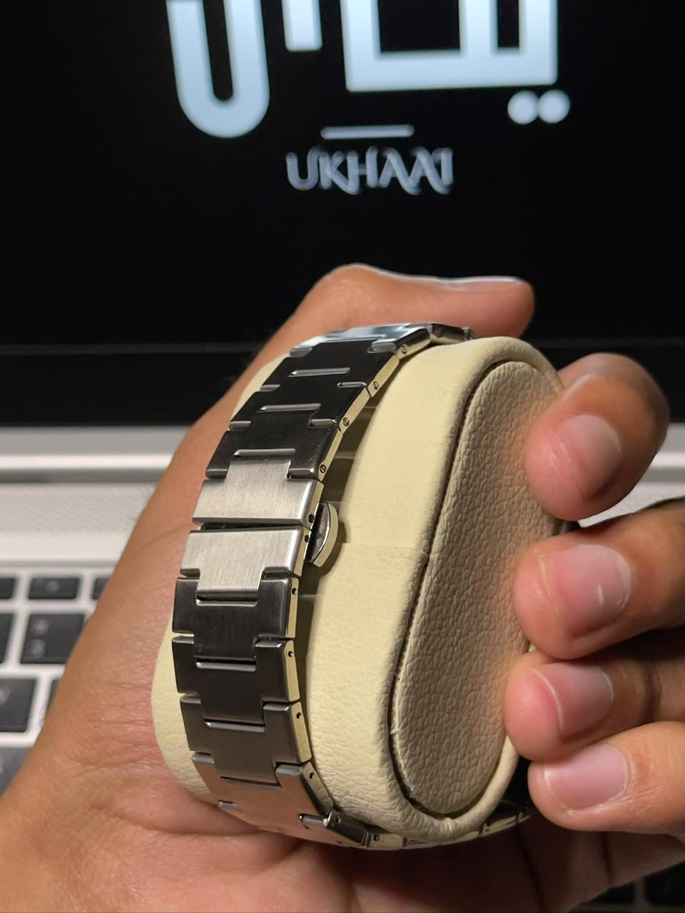 Hand holding a silver watch with a strap against a blurred background (back side)