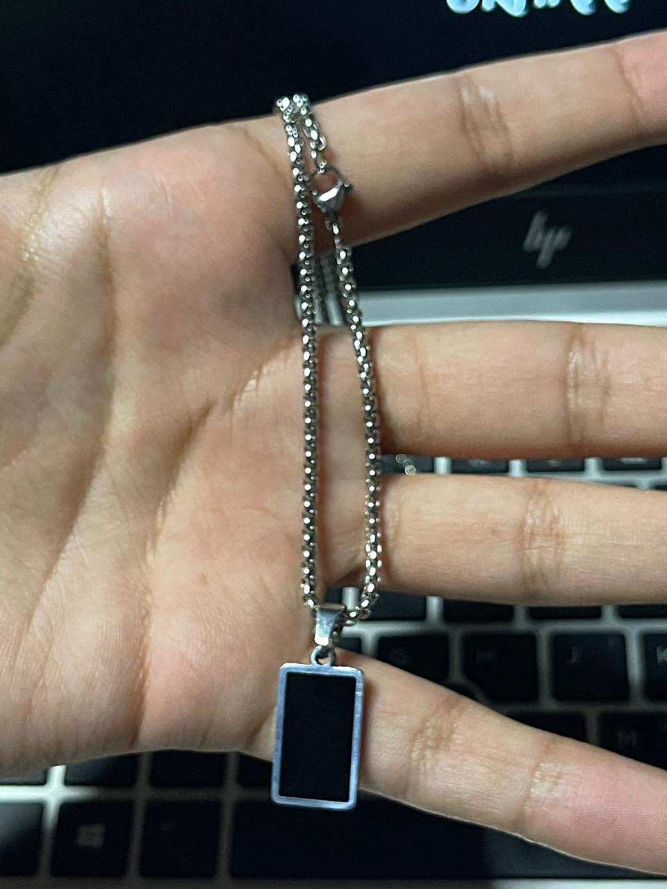 Hand holding a silver necklace with a rectangular pendant in front of a laptop front side.