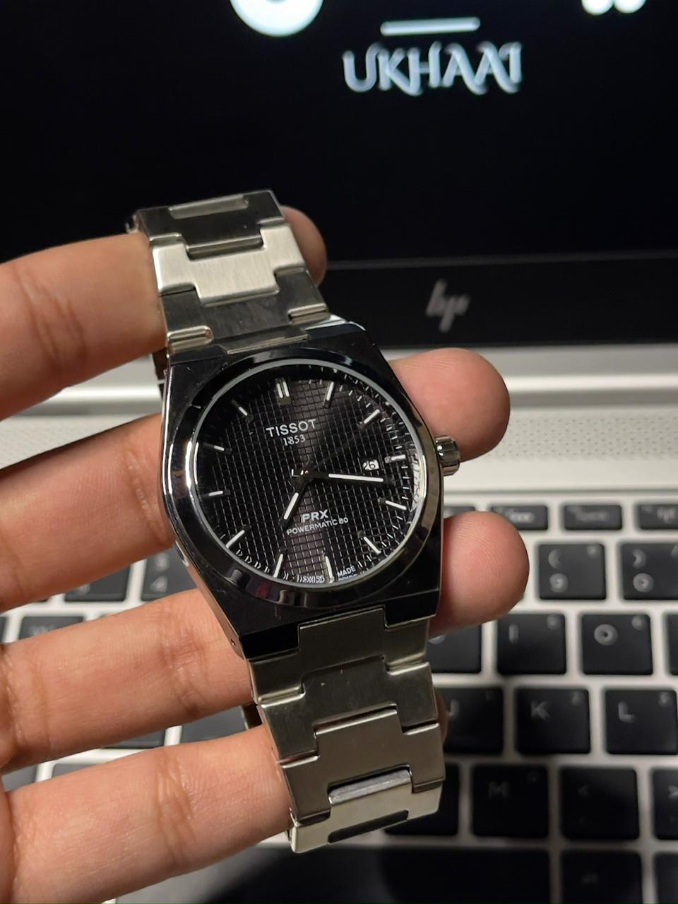 Silver Tissot watch held in a hand with a laptop in the background