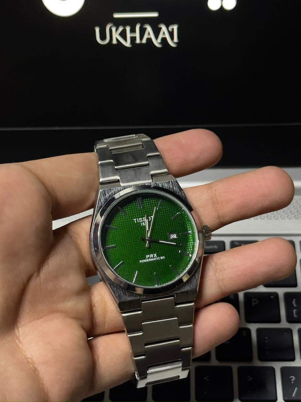 Hand holding a silver watch with a green face in front of a laptop with 'UKHAAT' branding.(front side)