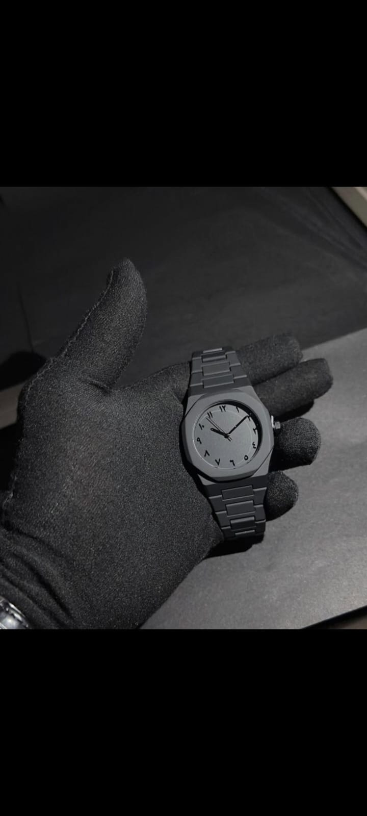 Hand wearing a dark glove holding a black dark wristwatch on a plain background front side.