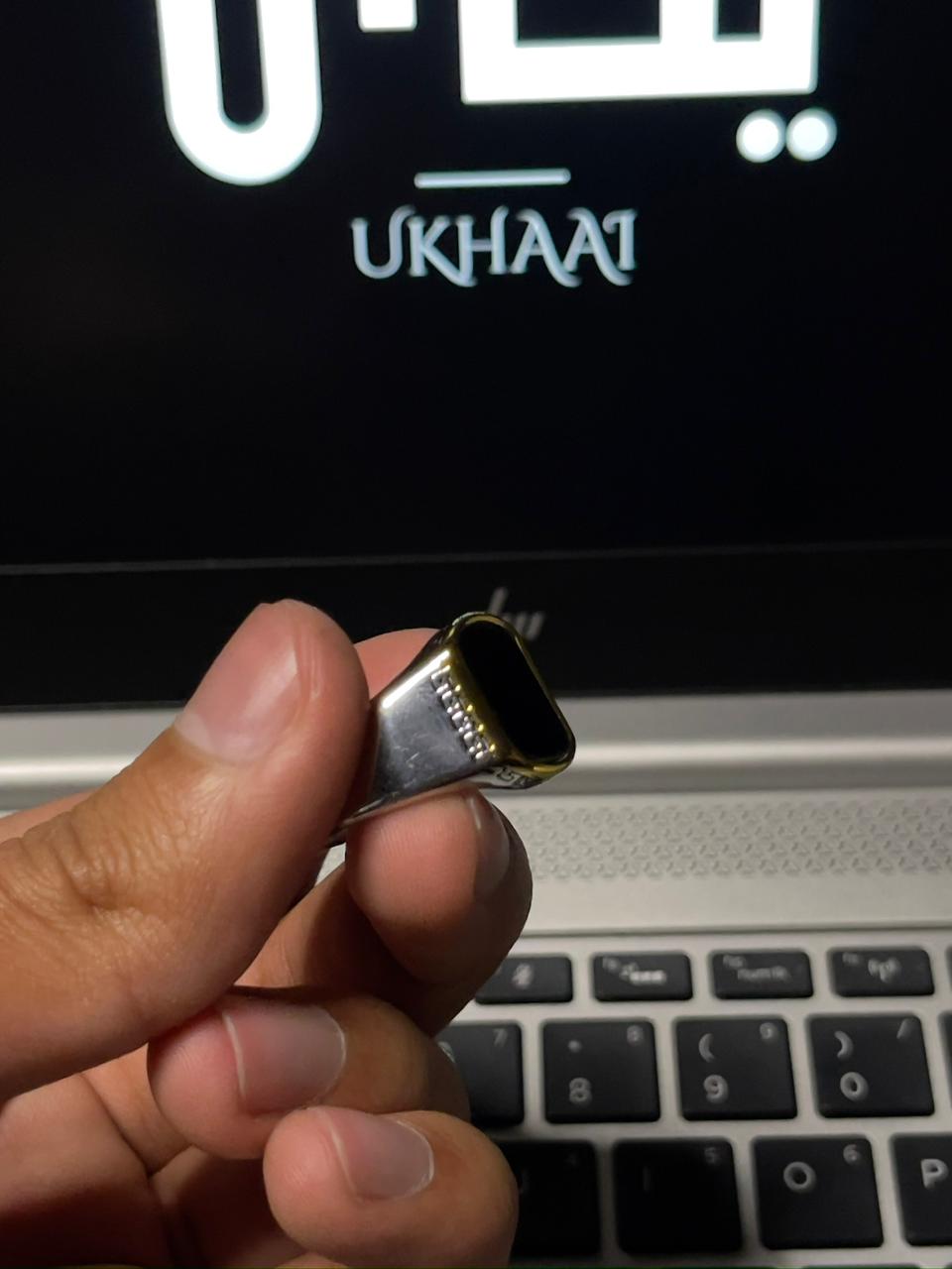 Hand holding a luxury ring   in front of a laptop with 'UKHAAI' branding.