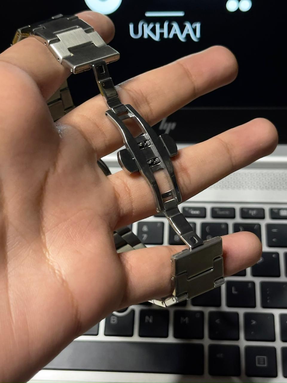 Metallic watch butterflylock held in a hand with a laptop in the background with 'Ukhaai' branding 